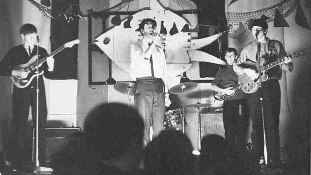 A vintage black and white image of a band performing on stage, featuring four male musicians playing guitars and singing, with a drum set and decorative backdrop.