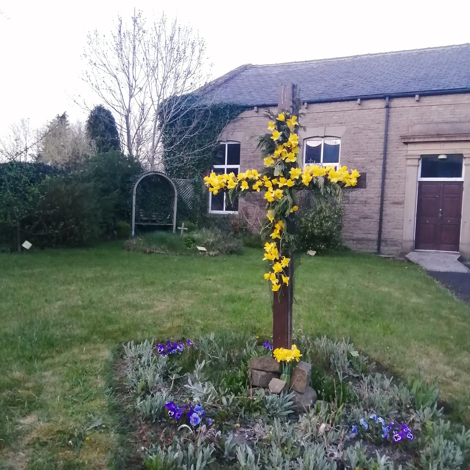 A wooden cross adorned with yellow daffodil flowers, surrounded by a circular flower bed with purple and blue pansies in a grassy area next to a building.