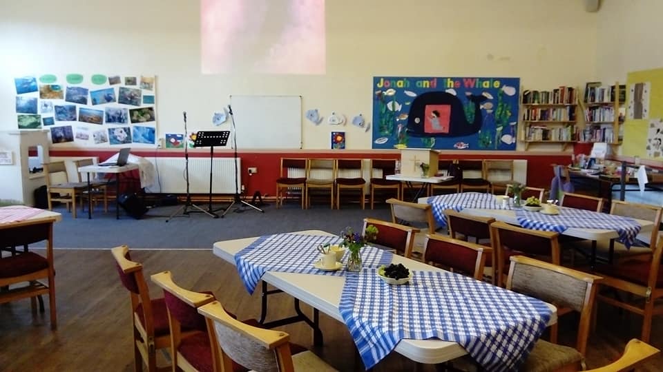 A vibrant community room with tables covered in blue checkered tablecloths, chairs arranged, a stage set up with a microphone, and colorful wall decorations including a whale and various pictures.