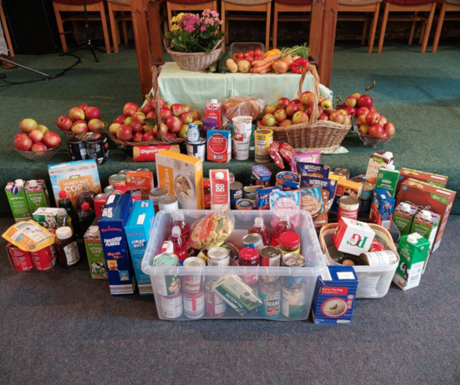 A collection of food items including apples, canned goods, and packages of various snacks displayed on a green carpet. All of the non-perishable items are for our local food bank.